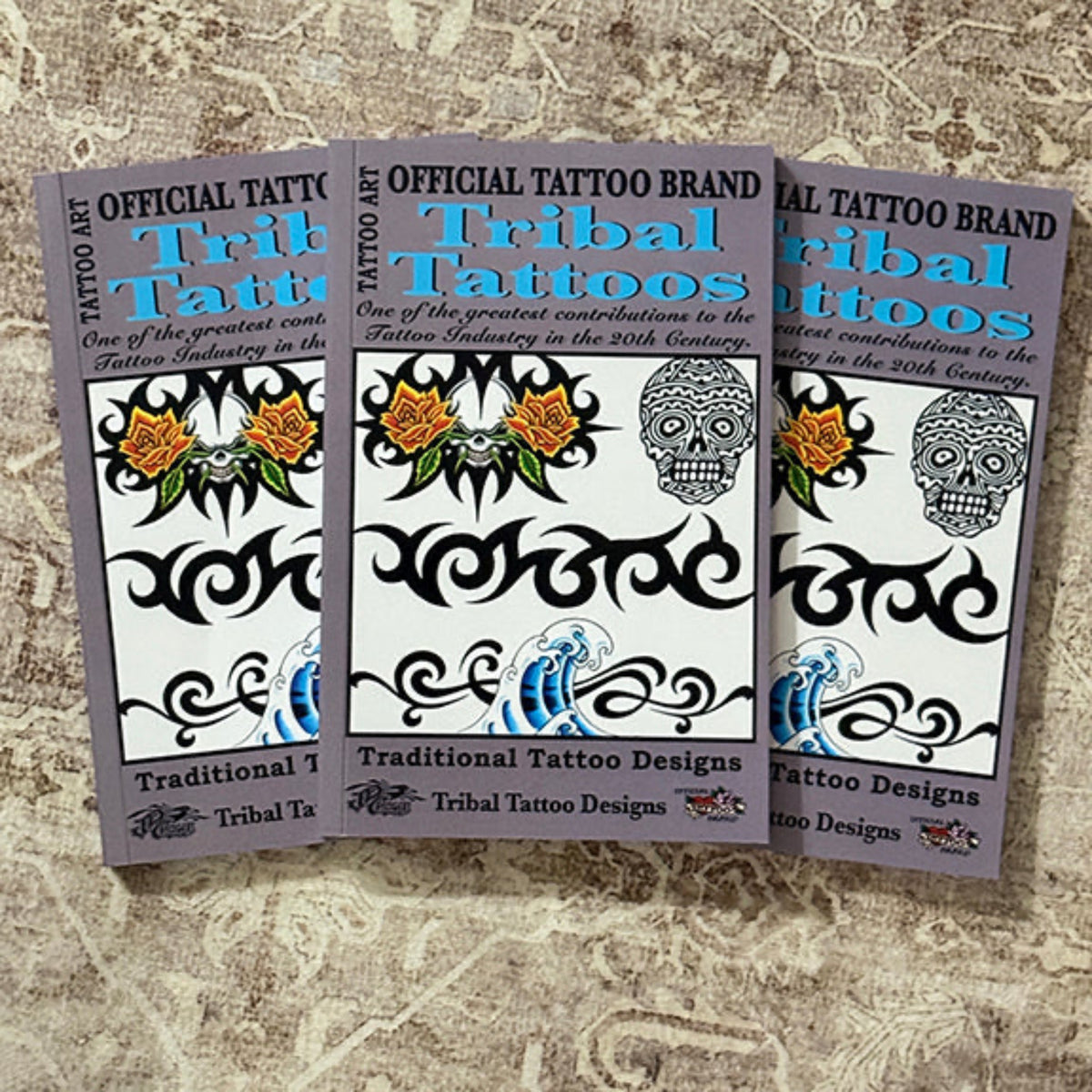 Three printed promotional cards or flyers for tribal tattoo designs are arranged overlapping on a textured beige fabric surface. The cards feature the text 'Official Tattoo Brand Tribal Tattoos' and display black tribal line art, floral motifs, a sugar skull, and blue wave designs.